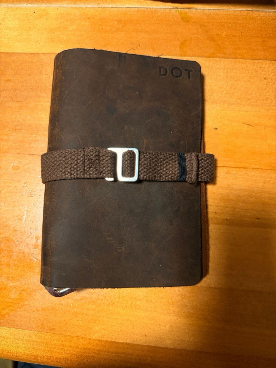 An early handmade DOT prototype: a brown leather notebook held closed with a woven strap and metal clip